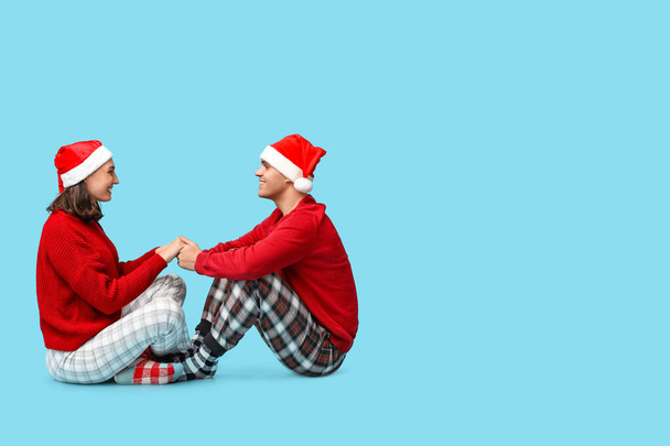 Mutlu genç çift, Noel pijamaları ve Noel Baba şapkaları ile mavi arka planda oturuyorlar. - Fotoğraf, Görsel