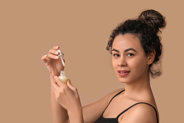 Kaunis nuori afroamerikkalainen nainen, jolla on pullo kosmeettista öljyä ruskealla taustalla - Valokuva, kuva