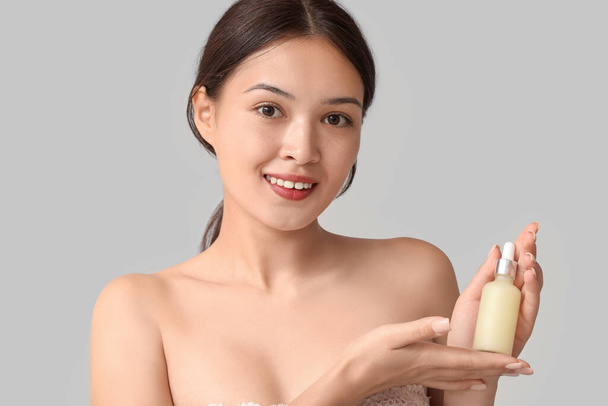 Kaunis nuori aasialainen nainen, jolla on pullo kosmeettista öljyä harmaalla taustalla - Valokuva, kuva