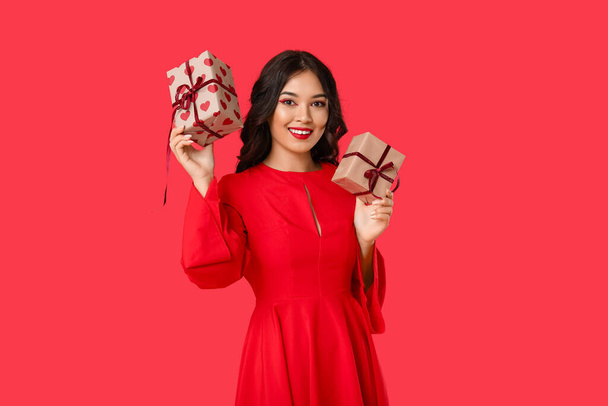 Όμορφη νεαρή ευτυχισμένη Ασιάτισσα γυναίκα με κουτιά δώρων σε κόκκινο φόντο. Γιορτή του Αγίου Βαλεντίνου - Φωτογραφία, εικόνα