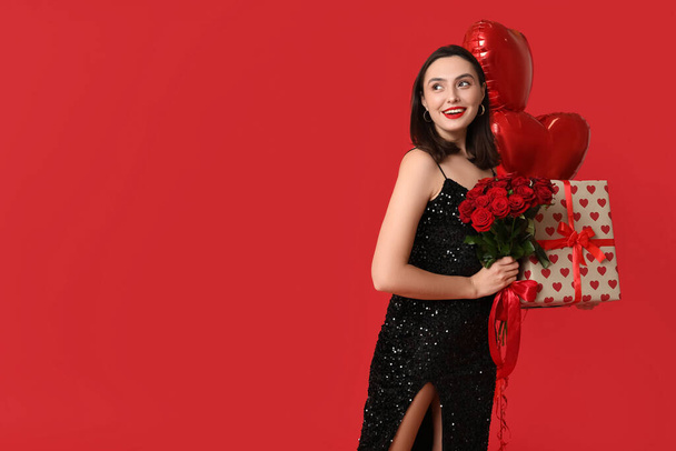 Όμορφη νεαρή ευτυχισμένη γυναίκα με κουτί δώρου, μπουκέτο τριαντάφυλλα και μπαλόνια σε σχήμα καρδιάς σε κόκκινο φόντο. Γιορτή του Αγίου Βαλεντίνου - Φωτογραφία, εικόνα
