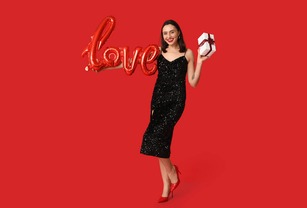 Beautiful young happy woman with gift box and balloon in shape of word LOVE on red background. Valentine's Day celebration - Photo, Image