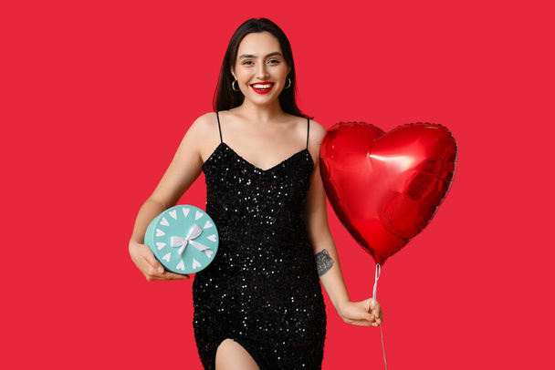 Gyönyörű, fiatal, boldog nő ajándékdobozzal és szív alakú lufival, piros háttérrel. Valentin-napi ünneplés - Fotó, kép