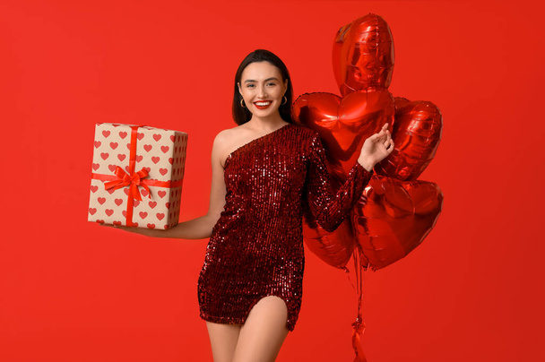 Belle jeune femme heureuse avec boîte cadeau et ballons en forme de coeur sur fond rouge. Fête de la Saint-Valentin - Photo, image