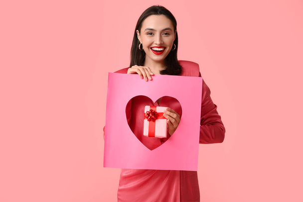 Beautiful young happy woman holding gift box and paper sheet with hole in shape of heart on pink background. Valentine's Day celebration - Photo, Image