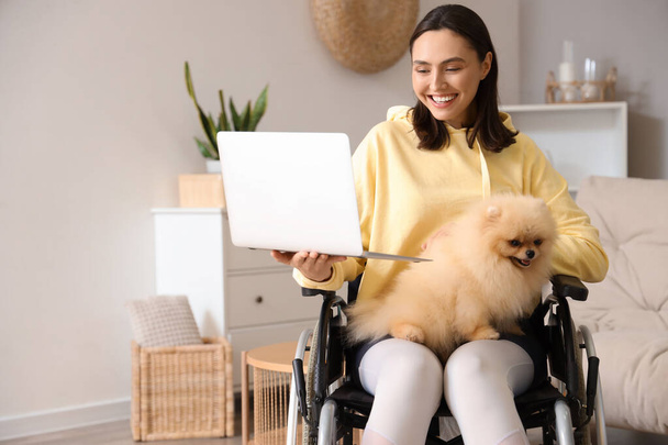 Mujer joven y deportiva en silla de ruedas con portátil y perro Pomeranian en casa - Foto, imagen