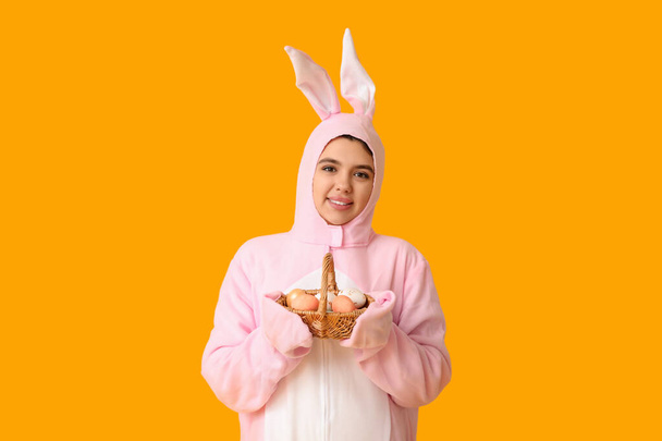 Krásná mladá žena v králičí kostým drží proutěný koš s velikonočními vejci na žlutém pozadí - Fotografie, Obrázek