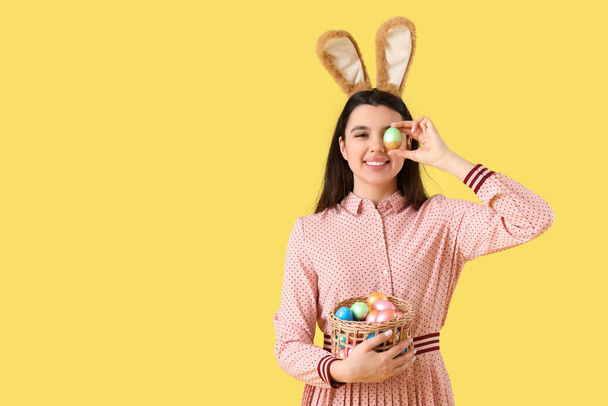 Beautiful young woman in bunny ears holding wicker basket with Easter eggs on yellow background - Photo, Image
