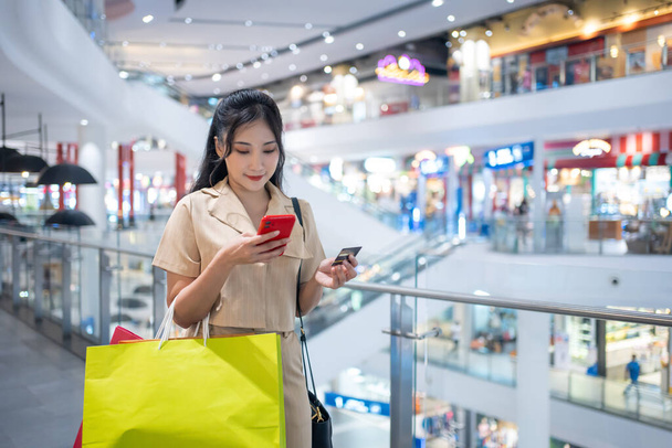 Gündelik giyinen genç bir kadın alışveriş merkezinde kredi kartıyla online alışveriş torbaları taşıyor. - Fotoğraf, Görsel