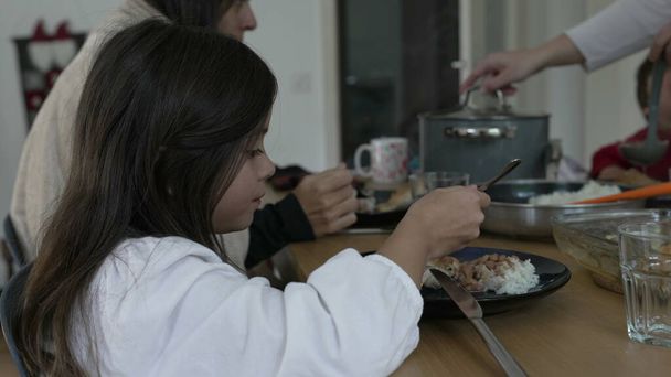 Сім'я насолоджується їжею разом, маленька дівчинка ріже їжу виделкою та ножем, спостерігаючи за динамікою сім'ї за обіднім столом, відвертим та автентичним домашнім способом життя - Фото, зображення