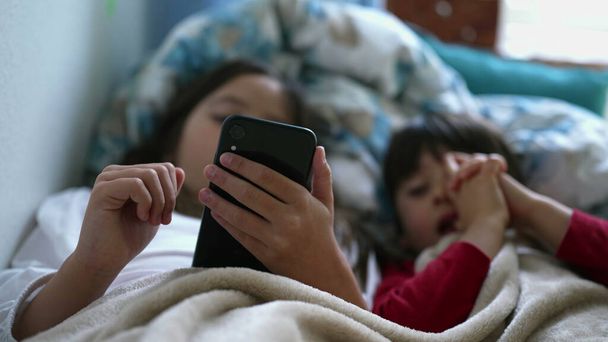 Küçük kızın eli yatak çarşaflarının altında cep telefonu cihazını tutarken kardeşi de yanında ekranı izliyordu. Modern teknolojiyle meşgul iki çocuk. - Fotoğraf, Görsel