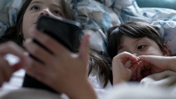 Çocuklar telefon ekranına bakıyor. Kız kardeş cep telefonu tutarken erkek kardeş internette medyaya bakıyor. Çocuklar yatak örtüsünün altında modern teknolojiyle meşgul. - Fotoğraf, Görsel
