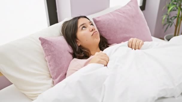 Mujer hispana joven despertando en un ambiente acogedor dormitorio interior, buscando somnoliento y cómodo bajo la ropa de cama blanca. - Metraje, vídeo