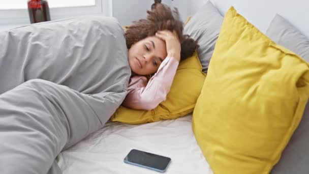 Junge hispanische Frau mit lockigem Haar checkt Telefon beim Aufwachen in ihrem gemütlichen, bunten Schlafzimmer. - Filmmaterial, Video