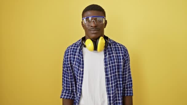 Selbstbewusster afrikanisch-amerikanischer Mann mit einem gesunden zahmen Lächeln, der in einer Schutzbrille steht und positiv, glücklich und freundlich aussieht. isoliert auf einem fröhlichen gelben Hintergrund. - Filmmaterial, Video