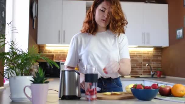 Mladá žena doma v kuchyni dělá ovocný koktejl. Krásná šťastná mladá žena v neformální domácí kuchyni se připravuje fitness koktejl doma. Sportovní a veganské jídlo - Záběry, video