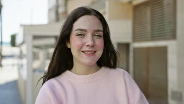 Egy fiatal nő rózsaszín pulóverben mosolyog a kamerába egy napos városi utcán.. - Felvétel, videó