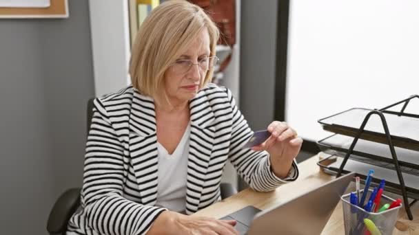 Starší žena zkoumá kreditní kartu skepticky v kanceláři stolu s notebookem a pery - Záběry, video