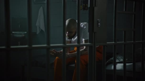 Criminal in orange uniform sits on prison cell bed. Prisoner serves imprisonment term for crime in jail. Gangster in detention center, correctional facility. Justice system. View through metal bars. - Footage, Video