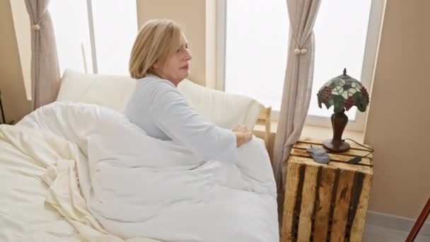 Vanhempi nainen pyjamassa venyttää selkänsä heräämisen jälkeen valoisassa makuuhuoneen sisustuksessa kotona. - Materiaali, video