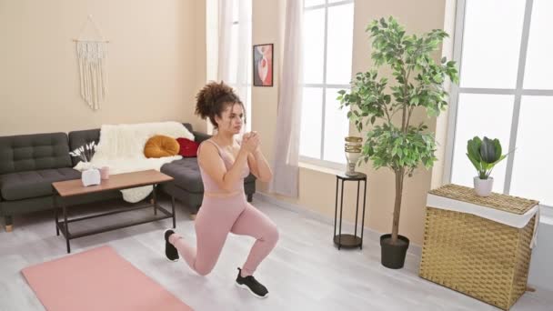 Una mujer joven con ejercicios de pelo rizado en una sala de estar bien iluminada, mostrando fitness y un estilo de vida saludable en casa. - Imágenes, Vídeo