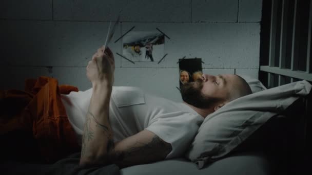 Narancssárga egyenruhás férfi fekszik az ágyon a cellában, képeket néz a családról és a gyerekekről. Illegálisan elítélt ember börtönben tölti a büntetését. Fogolytábor vagy büntetés-végrehajtási intézet. - Felvétel, videó