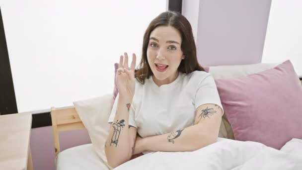 Wesoła młoda kobieta w piżamie liczy do trzech palcami, uśmiechając się jasno, doskonaląc komunikację gestów dłoni podczas relaksu w przytulnej sypialni - Materiał filmowy, wideo