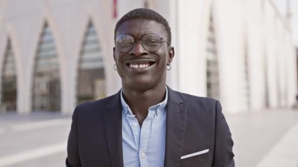 Hombre africano sonriente en traje de pie en la calle de la ciudad con arquitectura moderna en el fondo - Metraje, vídeo