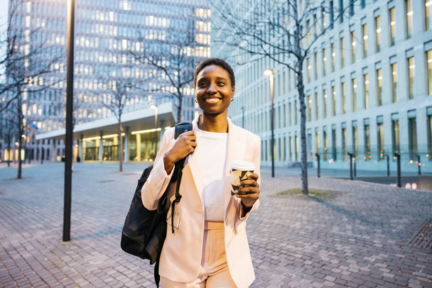Joven mujer de negocios hermosa con caminar en la calle con una mochila y un café. Empresaria africana que va a trabajar por la mañana. - Foto, Imagen