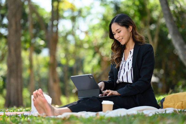 Ευτυχισμένη ξυπόλητη επιχειρηματίας που εργάζεται σε ψηφιακή ταμπλέτα, κάθεται στο γρασίδι στο πάρκο. - Φωτογραφία, εικόνα