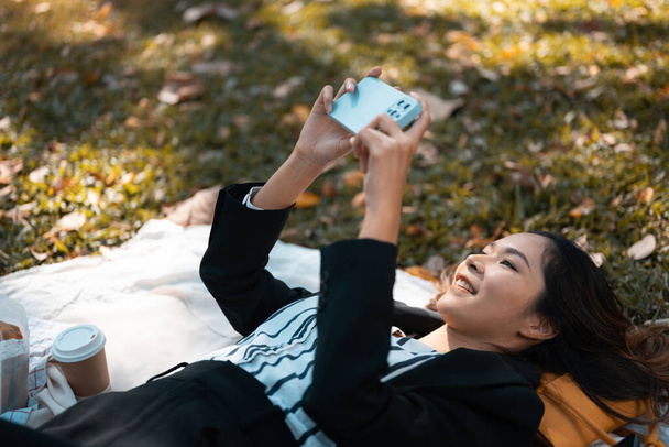 Onnellinen liikenainen makaa nurmikolla ja käyttää matkapuhelinta syksyllä puistossa. - Valokuva, kuva
