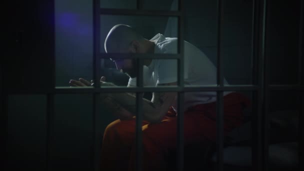 Narancssárga egyenruhás bűnöző ül az ágyon a cellában, feláll és rácsos ablakot néz. A rab börtönbüntetést kapott a börtönben elkövetett bűncselekményért. Gengszter a fogdában. Átlátszó fém rúd. - Felvétel, videó