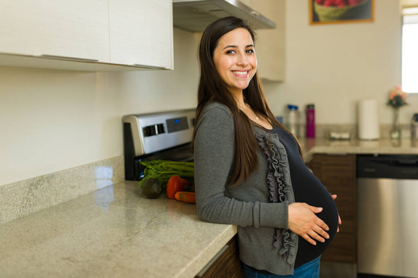 Upea raskaana oleva nainen hymyilee katsekontakti kotona keittiössä ajattelemalla vauvaansa ja äitiyttään - Valokuva, kuva