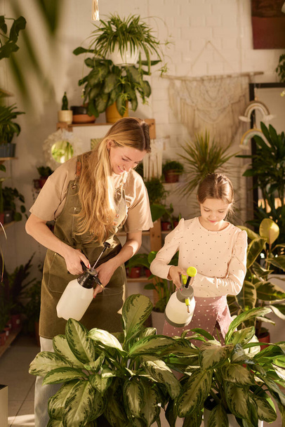 Вертикальный снимок девушки, помогающей своей веселой матери на работе в современном цехе, распыляющей воду на комнатные растения - Фото, изображение