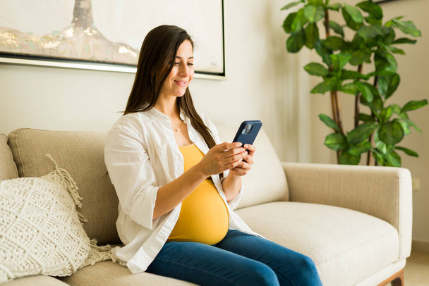 Mooie zwangere Kaukasische vrouw glimlachen tijdens het sms 'en op de smartphone terwijl ontspannen op de bank thuis  - Foto, afbeelding
