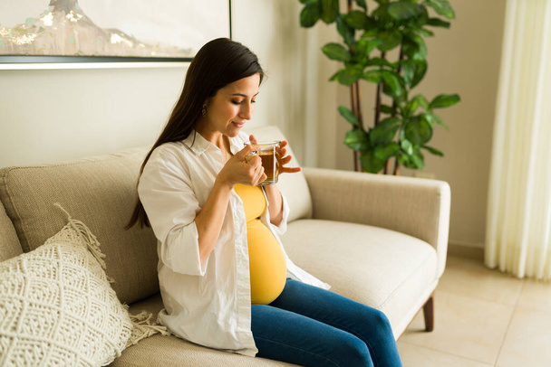 Веселая, роскошная беременная женщина улыбается, расслабляясь в гостиной, пьет чай и думает о своем ребенке - Фото, изображение