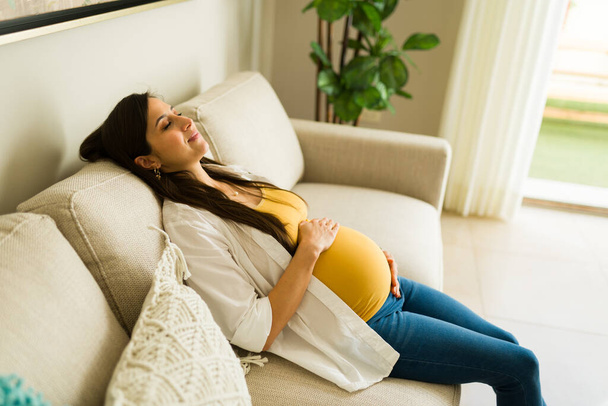 Mujer embarazada caucásica descansando en el sofá y relajándose en la sala de estar mientras se ve relajada disfrutando de su embarazo - Foto, Imagen