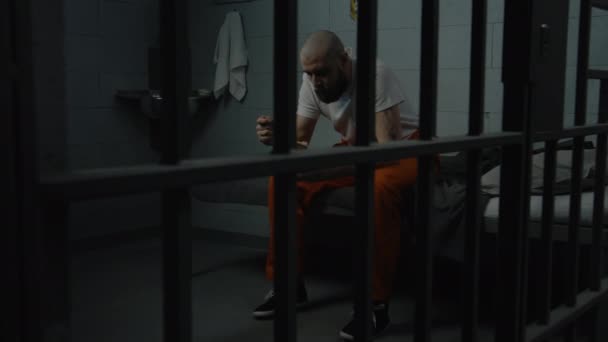Mannelijke gevangene in oranje uniform zit op bed in de cel en probeert walgelijk gevangenisvoedsel te eten uit een ijzeren kom. Crimineel zit gevangenisstraf uit. Detentiecentrum of correctionele inrichting. - Video