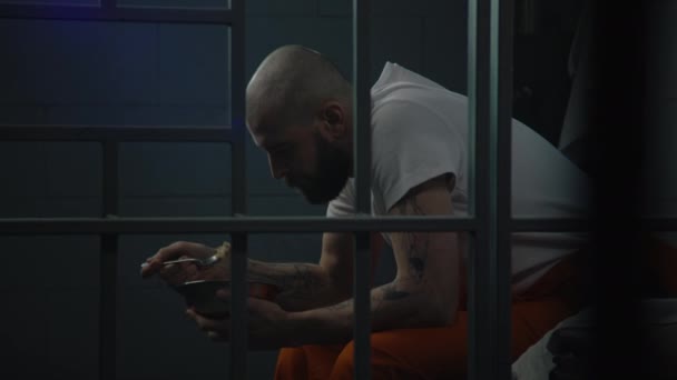 Мужчина в оранжевой форме сидит на кровати в тюремной камере, ест отвратительную тюремную еду из железной чаши. Заключенный отбывает тюремный срок за преступление в тюрьме. Исправительный центр или исправительное учреждение. - Кадры, видео