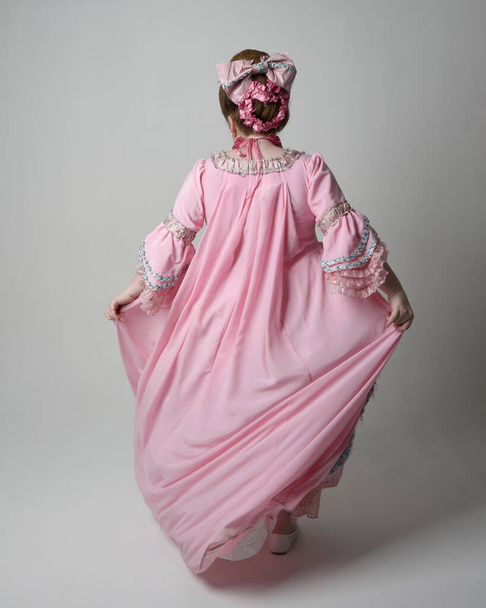 Full length portrait of woman wearing historical French baroque pink gown in style of Marie Antoinette with elegant hairstyle. Standing pose, walking away from the camera, isolated on studio background. - Photo, image