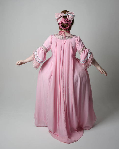 Plný portrét ženy v historických francouzských barokních růžových šatech ve stylu Marie Antoinette s elegantním účesem. Stojící póza, vzdalující se od kamery, izolované na pozadí studia. - Fotografie, Obrázek