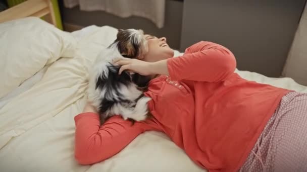 Een jonge vrouw ontspant op een bed terwijl ze liefdevol haar biewer yorkshire terriër aaien in een gezellige slaapkamer setting. - Video