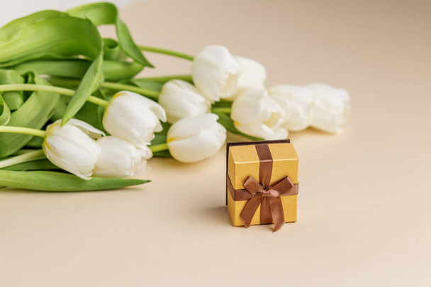 Composition de bon goût avec des tulipes blanches à côté d'une charmante boîte cadeau avec un ruban brun, sur fond beige neutre. - Photo, image