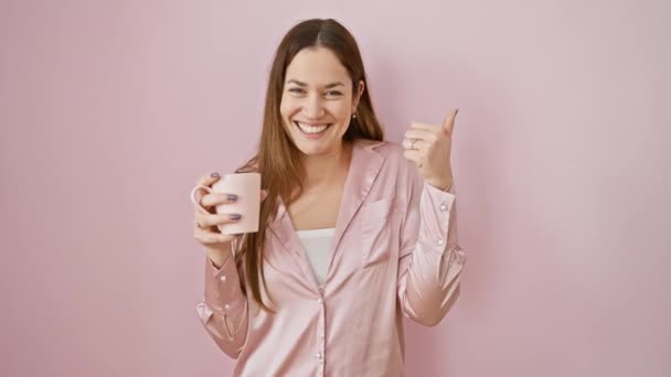 Opgewonden jonge vrouw met blauwe ogen, vrolijk koffiekopje vasthoudend, zelfverzekerd duim naar de zijkant wijzend terwijl ze een pyjama draagt, vrolijk lachend met open mond over geïsoleerde roze achtergrond. - Video