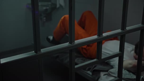 Inmate in orange uniform lies on prison cell bed. Prisoner serves imprisonment term for crime. Criminal in detention center, correctional facility. Justice system. View through metal bars. Dolly shot. - Footage, Video