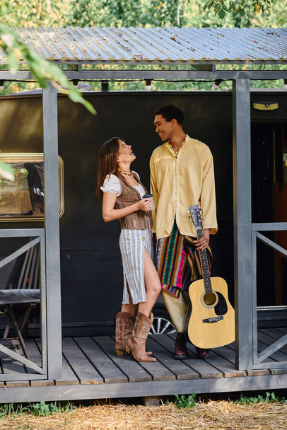 Ένα διαφυλετικό ζευγάρι, άντρας και γυναίκα, που στέκονται μαζί με μια κιθάρα στο χέρι, και τα δύο περιβάλλονται από τη φύση.. - Φωτογραφία, εικόνα