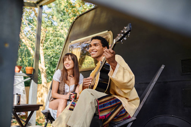 Egy férfi gitározik, miközben egy nő mellett ül, harmonikus dallamot hoz létre a tábortűz mellett, természetes környezetben.. - Fotó, kép