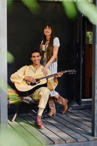 Ένας άντρας και μια γυναίκα κάθονται σε μια βεράντα παίζοντας κιθάρα μαζί, απολαμβάνοντας μια στιγμή μουσικής σύνδεσης σε ένα γαλήνιο υπαίθριο περιβάλλον. - Φωτογραφία, εικόνα