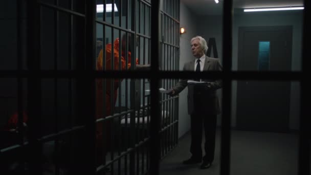 Crimineel in oranje uniform leunt op cel bars, praat met advocaat en leest zijn contract. Gevangene zit gevangenisstraf uit voor misdaad in de gevangenis. Gangster in detentiecentrum. - Video
