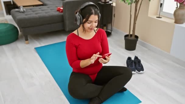 Una joven hispana con un top rojo usa un teléfono inteligente mientras está sentada en una alfombra azul en casa, usando auriculares. - Metraje, vídeo